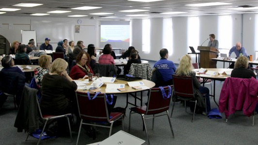 A large room of people sitting at tables in a UFCW 832 Training Centre classroom, listening to a speaker at a podium, for a teaching session or meeting.