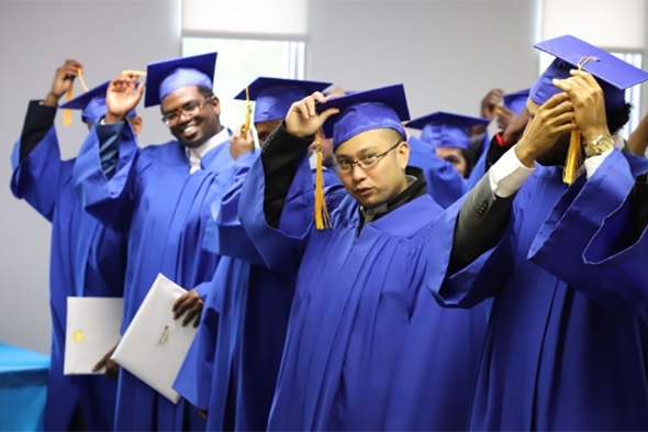 Several adult high school graduates, in their blue graduation robes, standing holding their graduation caps, getting ready to turn over their golden tassels. 