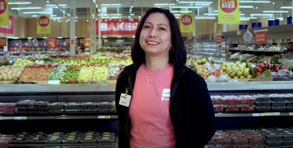 A smiling female grocery worker, with black hair, wearing a black sweater and a pink t-shirt, stands in the produce department.