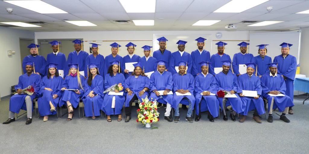 A graduation photo with large group of adult students, wearing blue cap and gowns, posing for a formal photo in a classroom.