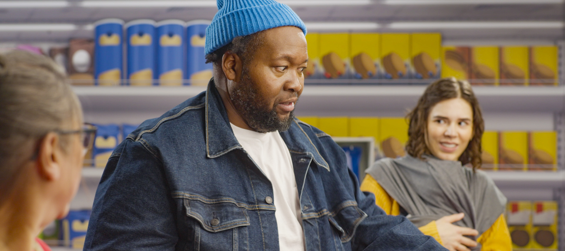 A middle-aged man wearing a denim jacket, white tee shirt and blue tuque looks confused at a self-checkout machine in a grocery store as an older woman and a younger woman holding a baby look up at him from the self-checkout machines they are using.