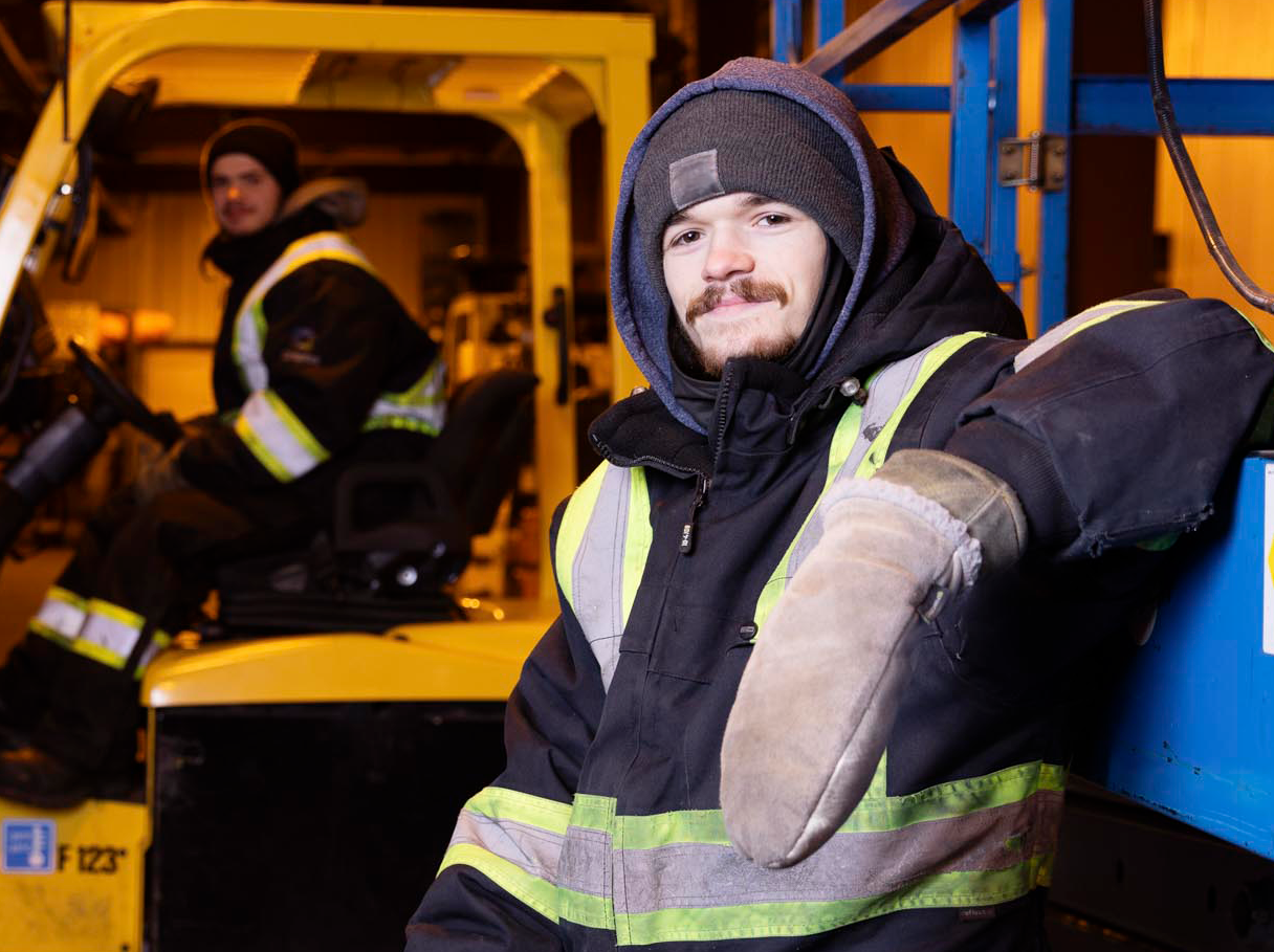 A ufcw member leaning against some machinery while another ufcw member is in the background on a forklift.